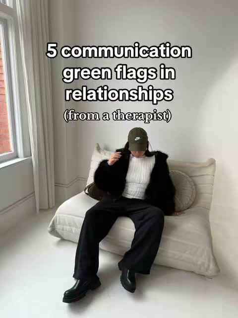 A well-lit interior shot features a person seated on a large, cream-colored cushion. The individual, dressed in a black jacket, white top, and dark pants, is wearing a baseball cap and black shoes. Text overlayed on the image reads '5 communication green flags in relationships (from a therapist)'. The background includes a bright window and minimal decor.