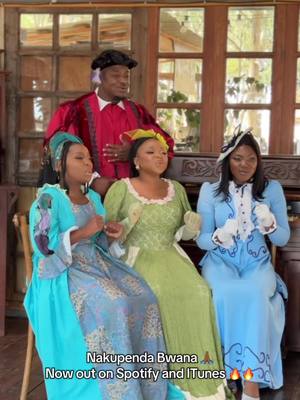 An image capturing a group of four people, likely performers, dressed in vintage-style attire. The setting appears to be outdoors, possibly a stage or an area with wooden accents and large windows. Text overlays indicate the name of a song and where it can be found.