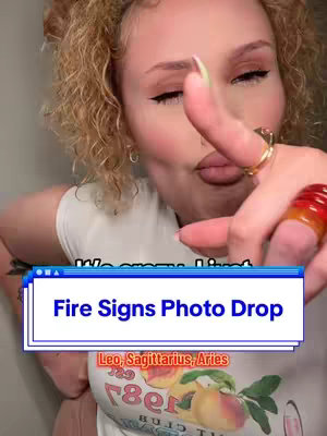 A woman with light skin, curly blonde hair and visible makeup is shown in a selfie-style shot. She is pointing at the camera with her index finger. A text overlay reads "Fire Signs Photo Drop" with the subtext, "Leo, Sagittarius, Aries". She is wearing a white t-shirt with a colorful print. The background is neutral.