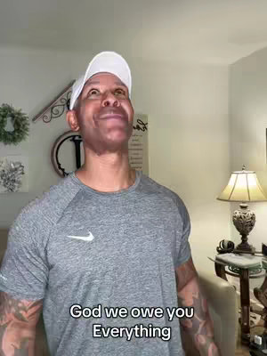 A man wearing a white cap and a grey t-shirt is shown looking upwards with an expression of reverence. Below him is overlaid text with the message 'God we owe you Everything'. The background is a simple domestic setting with light-colored walls and a lamp in the background.