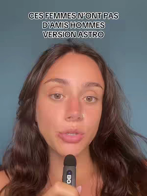A woman with long brown hair is centered in the frame; she's holding a microphone and looking directly at the viewer. Above her, there's text overlay in a bold, sans-serif font. The backdrop behind her is a simple, light blue wall.