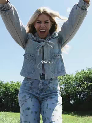 A woman with long blonde hair stands outdoors on a sunny day, arms raised in a joyful gesture. She wears a light blue denim jacket and matching jeans. The background includes green trees and a clear blue sky with a text overlay reading "O Lord".