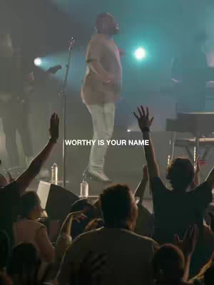 A live performance on stage with a singer in the spotlight, arms raised, and a crowd beneath them. Light shines down on the stage, and the text 'WORTHY IS YOUR NAME' is overlaid.