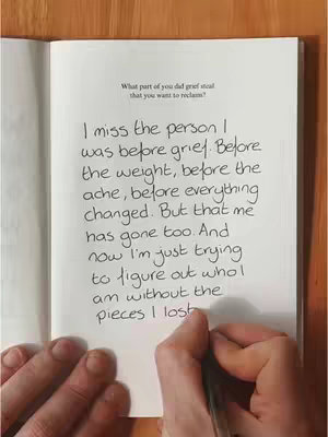 The image shows an open notebook on a wooden surface, with handwritten text filling most of the right page. A hand holding a pen is visible, in the process of writing. The text starts with a question and presents a personal reflection on grief.