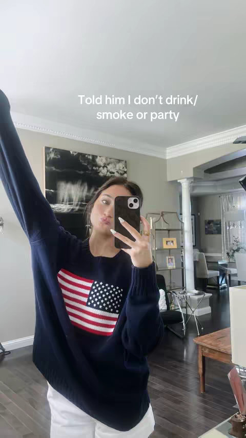 A woman takes a selfie, holding her phone in front of her face. She is wearing a navy blue sweater with a large American flag design on the front. Her arms are raised, and she appears to be in her home, as a mirror and other home decor can be seen in the background.