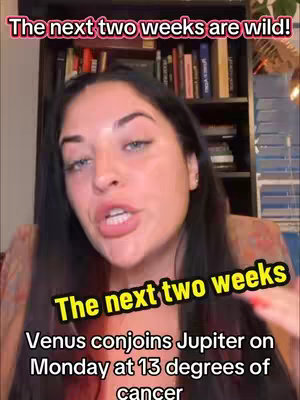 A woman with long dark hair is centered in the frame, speaking directly to the viewer, the background blurred out but contains books. Overlayed text includes a banner at the top reading 'The next two weeks are wild!' and an additional text overlay at the bottom stating 'The next two weeks' , with additional information. The lighting appears warm, suggesting an indoor setting.