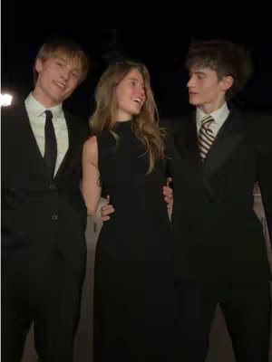 Three young people, two men flanking a woman, pose together in what appears to be a formal setting. All three are dressed in black suits. The woman is positioned in the center, wearing a sleeveless black dress, and is smiling and looking towards her left. The man on her right has a striped tie and smiles back at her, his arm around her waist. The man on her left has a black tie and is also smiling, leaning slightly towards her. Dark background.