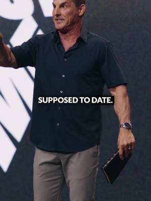 A man in a dark blue button-down shirt and khaki pants is standing on a stage, gesturing with one arm. Text overlayed reads 'SUPPOSED TO DATE.' He holds a black book in his other hand. The background is blurred, suggesting a stage setting with dark tones.