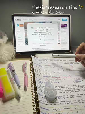 A desk setup with a laptop displaying a research paper, surrounded by pens, highlighters, and a notebook filled with notes. The image has a warm, inviting colour palette with good lighting.