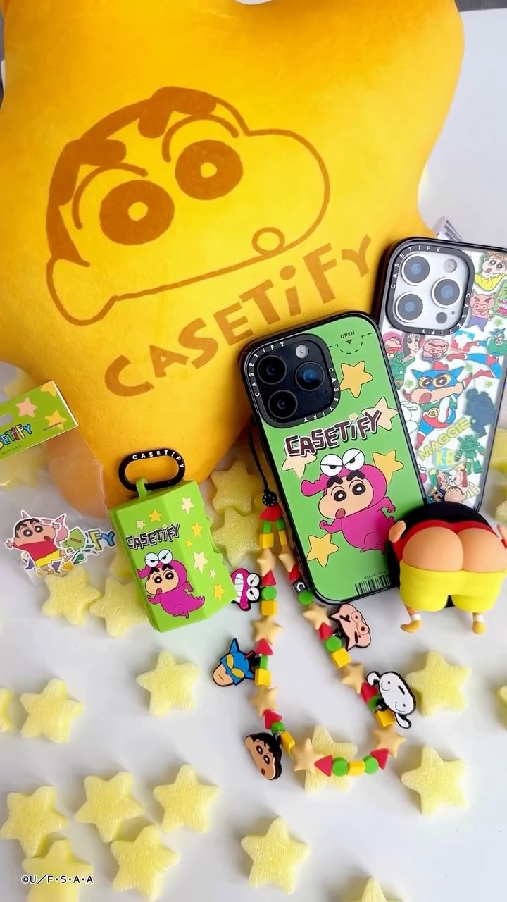 The image displays a collection of Crayon Shinchan-themed merchandise arranged on a white surface. Items include a yellow plush pillow with Shinchan's face, iPhone cases, an AirPod case, and a beaded charm necklace with Shinchan and other character charms. Star-shaped foam pieces are scattered around the items, adding to the playful theme. The overall composition is bright, colorful, and visually appealing.