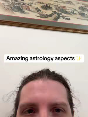 The video features a selfie-style shot with the creator's face in the foreground. Above the creator, a framed piece of artwork is visible on the wall. The text overlay reads 'Amazing astrology aspects ✨.' The background is a plain white wall. The overall composition is simple and direct, focusing on the creator and the overlay text.