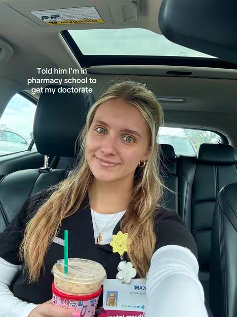 A young woman with blonde highlights is seated inside a car. She is wearing a black uniform, possibly work attire, and has a name tag and ID pinned to her chest. She holds a cup of iced coffee in one hand. Text overlayed on the image reads "Told him I'm in pharmacy school to get my doctorate."