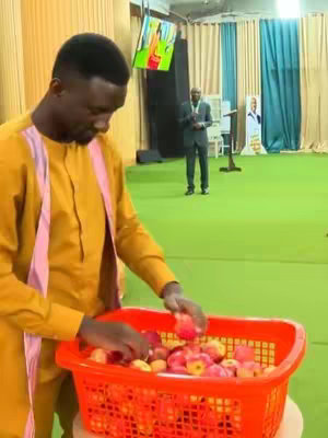 A man in a yellow shirt and pink scarf is shown in an indoor setting, possibly a stage or church. He is interacting with a large orange basket full of red apples. In the background, a person in suit stands, facing audience with a poster that has a person's art on the side.