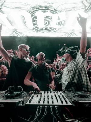 The image captures a dynamic music event, featuring three individuals centered around a DJ mixing console. Two are actively engaged, with arms raised, while a third individual stands amidst a crowd, all illuminated by a bright stage backdrop. The lighting suggests a night-time outdoor setting, punctuated by vibrant energy.
