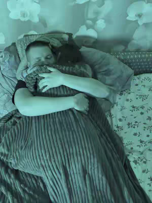 A man and a person are embracing in bed. The man is on the bed under a blanket that is wrapped around the other person and him.