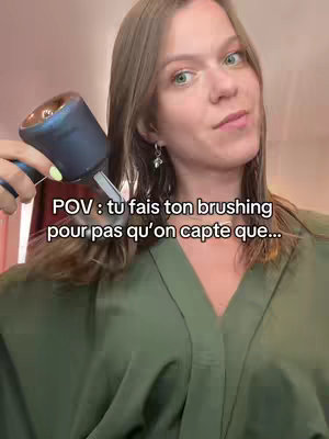 A woman is featured in a selfie-style video with text overlayed. The woman is holding a hair styling tool near her face and wearing a green jacket.