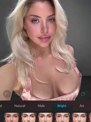 A woman with long blonde hair and green eyes is the focal point of a selfie video. She's wearing a pink top, and the lighting appears to be bright. The background is a blurred neutral tone. The bottom of the screen shows icons, suggesting some sort of app interface.