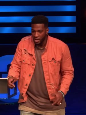 A man in an orange jacket and brown shirt is speaking, standing center-frame. The backdrop is a blurred blue gradient suggestive of a stage setting.