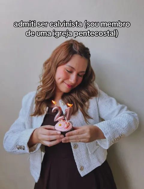 A woman holding a cupcake with candles in the shape of "21" in front of her. She has light brown hair, is smiling, and wearing a white blazer over a brown top. The background is a plain, light-colored wall.