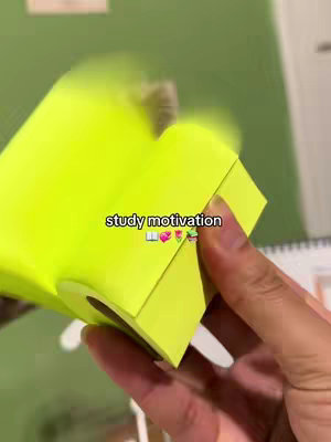 A person's hand holding a bright yellow, heart-shaped container with a cut-out. The container has a slot for the content to be removed from the side. The background is green and has other stationery and a white notebook in the background, with the text 'study motivation' on a white text box, with emojis.