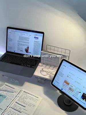 The image shows a well-lit workspace with a laptop, an iPad on a stand, and a notebook open on a desk. The laptop is open, displaying a document or webpage. The iPad is angled towards the viewer, showing an app interface. A pen and glasses are on the desk, next to a notebook displaying written notes. The overall composition presents a study or work setup, with cool-toned lighting.