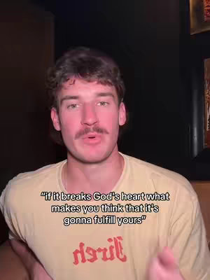 A man with short, dark hair and a mustache is centered in the frame, looking directly at the viewer. He's wearing a light-colored t-shirt with text on it and is positioned against a dark background. Overlayed on the image, in a sans-serif font, is the text:
"if it breaks God's heart what makes you think that it's gonna fulfill yours".