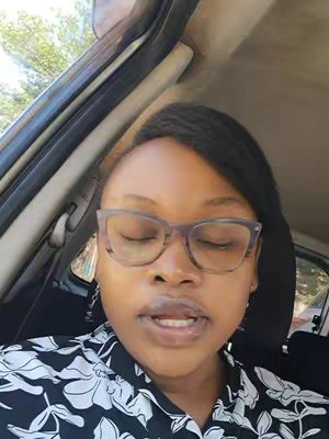 A woman with glasses is seated in a car, looking towards the camera with her eyes closed and mouth slightly open. She is wearing a black and white floral print shirt. The background shows a car interior, with glimpses of the outside world visible through the window.