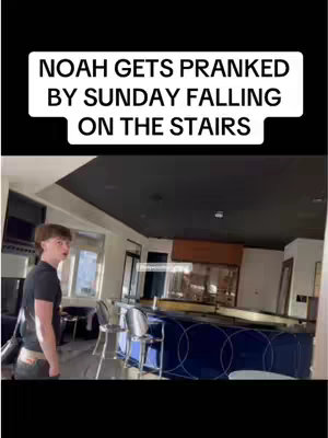 A young man stands in a well-lit, modern kitchen with a dark ceiling. Above the man, text overlaid on the image indicates a prank related to stairs. Interior includes a bar-like structure, and appliances. The overall composition suggests a vlog or comedic setup, filmed in what appears to be a home environment.
