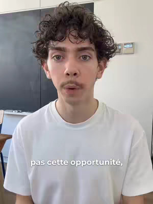A young man with curly brown hair and a mustache looks directly at the camera. He's wearing a white t-shirt, and a black board and some doors are visible in the background.