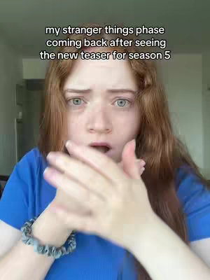A young woman with red hair is looking at the camera with wide eyes and a surprised expression. She is wearing a blue top and holding her hands up near her face, as if she is about to clap or cover her mouth.
