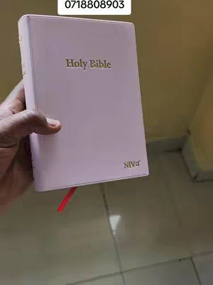 A hand holds a light pink book, likely a Bible, against a blurry yellow background. The book has gold lettering that says 'Holy Bible' on it. A red ribbon sticks out from the top. The hand holding the book is of a darker skin tone. Above the book, there is an additional rectangular shape with a number in it.