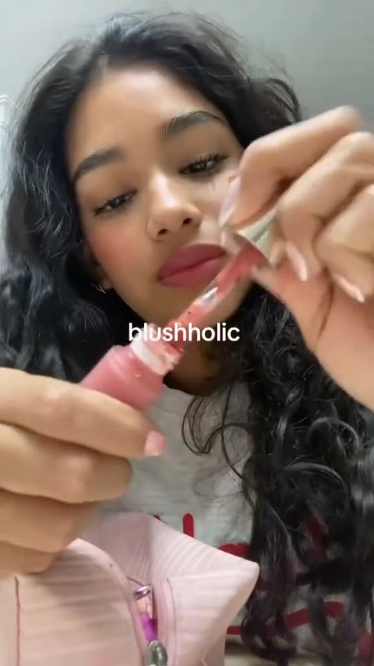 A selfie-style video shows a woman holding a tube of blush, squeezing it out. The woman has dark curly hair and is wearing a light-colored shirt. The word "blushholic" is shown in white letters at the bottom center of the frame.