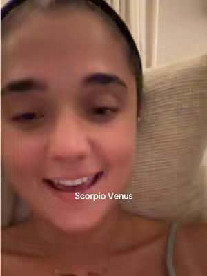 A close-up selfie shot of a young woman with dark hair and a black headband. She is sitting inside, and the background includes a neutral-colored pillow. "Scorpio Venus" is overlaid on the image in white, centered below her chin. The woman is smiling, and her expression is warm and inviting.