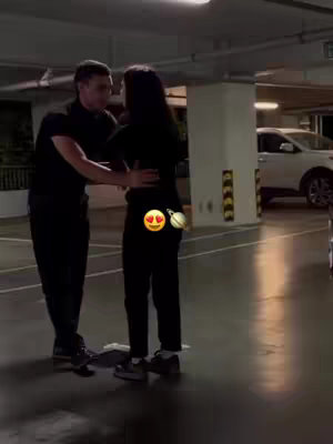 Two people, a man in a black shirt and pants, and a woman wearing a black top and pants, are embracing in a parking garage. The man is facing towards the woman and has his arms wrapped around her, while she has her arms around his torso. In the background, a part of a white car and a white pillar are visible. The image gives a sense of intimacy and affection.
