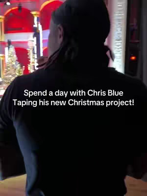 A man wearing a dark shirt and a cap is seen from the back, facing forward. Text overlayed on the image reads 'Spend a day with Chris Blue Taping his new Christmas project!' The background suggests a stage or studio setting with Christmas decorations.