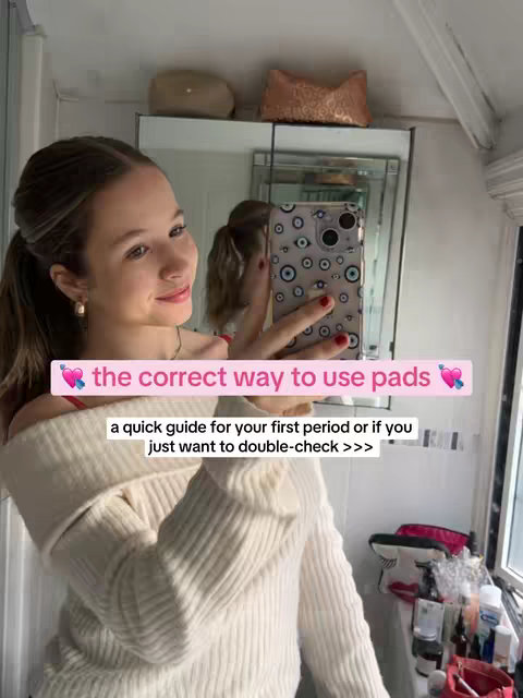 A selfie-style shot taken in a bathroom. A young woman stands facing the mirror, holding a phone in her hand to take a photo, with text overlays on the image. The woman's reflection is visible in the mirror. She is fair-skinned, with dark hair pulled back into a semi-ponytail and wearing a white sweater. A text overlay at the top of the image states, 'the correct way to use pads' in pink box. A second text overlay at the bottom of the image. The bathroom has a white ceiling and a mirror behind the woman. The overall composition is bright and well-lit.
