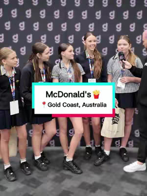 A group of six young women in school uniforms are being interviewed on the street. A graphic overlay indicates that the interview is about McDonald's and takes place on the Gold Coast, Australia, denoted by a location pin. The background is a wall with a repeating 'g' pattern.
