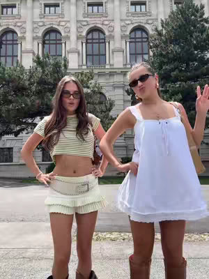 Two young women pose in front of a classical architectural building. They both wear sunglasses. One is wearing a crop top and shorts, while the other is wearing a white babydoll dress. The building is a light beige, with arched windows.