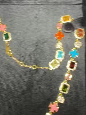 The image displays a gold necklace with various colorful gemstone charms on a dark gray or black background. The necklace is the central focus and takes up a large portion of the frame. The layout is simple, emphasizing the jewelry.
