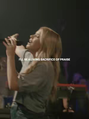 The image features a woman singing into a microphone on stage. Text overlayed on the image reads 'I'LL BE A LIVING SACRIFICE OF PRAISE'. The background shows a dimly lit stage with instruments visible.