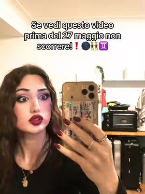 A selfie video cover shows a young woman holding her phone to record. She has brown hair, fair skin, and is wearing red lipstick and dark eye makeup. The text overlay says 'Se vedi questo video prima del 27 maggio non scorrere!'. Behind her is a room with a blurred background including a small oven, a suitcase and some plants.