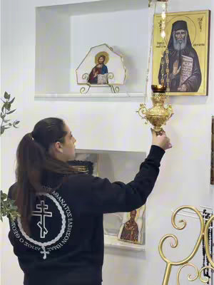 A woman holding a decorated gold lamp in front of a white wall. Behind her, there's a wall niche with religious icons and paintings. She's wearing a black hoodie with white text at the back. The overall setting implies a religious or spiritual theme.
