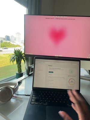 A well-lit indoor shot featuring a desk setup with a laptop, external monitor, and accessories. A window offers a view of the city skyline.  The monitor displays a pink background with a heart-shaped blur and text regarding Mendel's Law. The laptop screen has an open dashboard with visible elements. A hand is partially visible in the foreground, presumably of the creator. Various items are arranged on  the desk: headphones, a small plant in a pot.