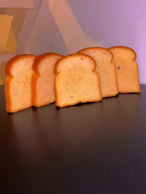 A close-up shot of five pieces of toasted bread arranged in a row on a dark surface, against a blurred background of a white structure and purple wall. The bread slices have a slightly golden hue with charred edges.