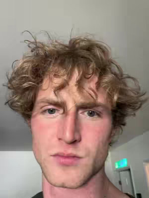 A close-up shot of a fair-skinned man with curly blonde hair and light eyes. His expression is neutral, and he is looking at the camera. The background is a plain, light-colored wall.