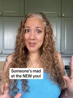 A woman with long, curly brown and gray hair is centered in the frame. She is wearing a blue top and a gold necklace, and her expression is animated, with wide eyes and a slightly open mouth. The background shows gray kitchen cabinets.