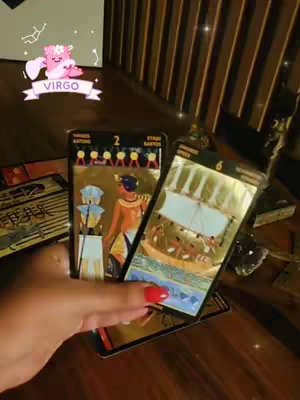 The image shows a person's hand holding up a tarot card. The cards depict Egyptian themed imagery with gold detailing. In the background, a table and decorative items can be seen and a banner with the text "VIRGO" is present.