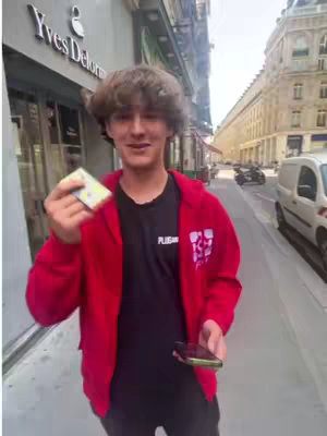 A young man stands on a sidewalk in front of a store with the sign 'Yves Delorm'. He holds a card in one hand and a smartphone in the other, smiling at the camera. He wears a red hoodie over a black t-shirt.