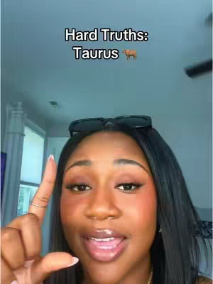 A close-up shot features a Black woman speaking into the camera, pointing with her index finger. Text overlay at the top reads "Hard Truths: Taurus" with a Taurus emoji next to it. The background shows a light-colored ceiling and a portion of a wall with a window.