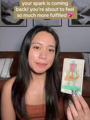 A woman with long black hair holds up a tarot card in front of her as she looks at the camera. Above her, the text overlay reads: "your spark is coming back! you're about to feel so much more fulfilled 💖". The image background includes framed artwork and a brown leather sofa.